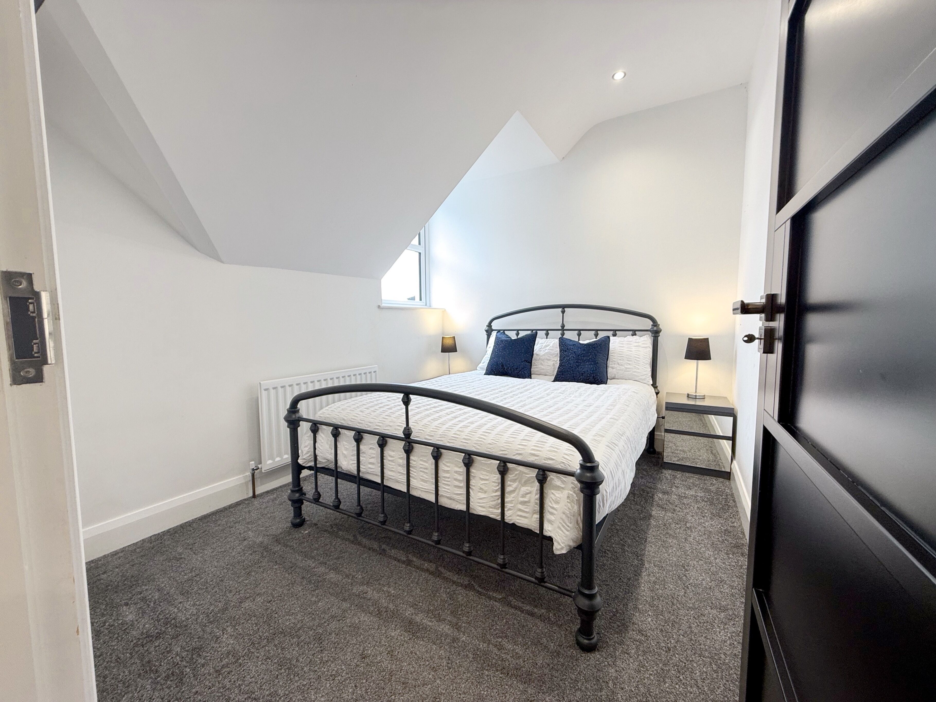 A bedroom with a bed, two bedside lamps, a radiator, and a window.