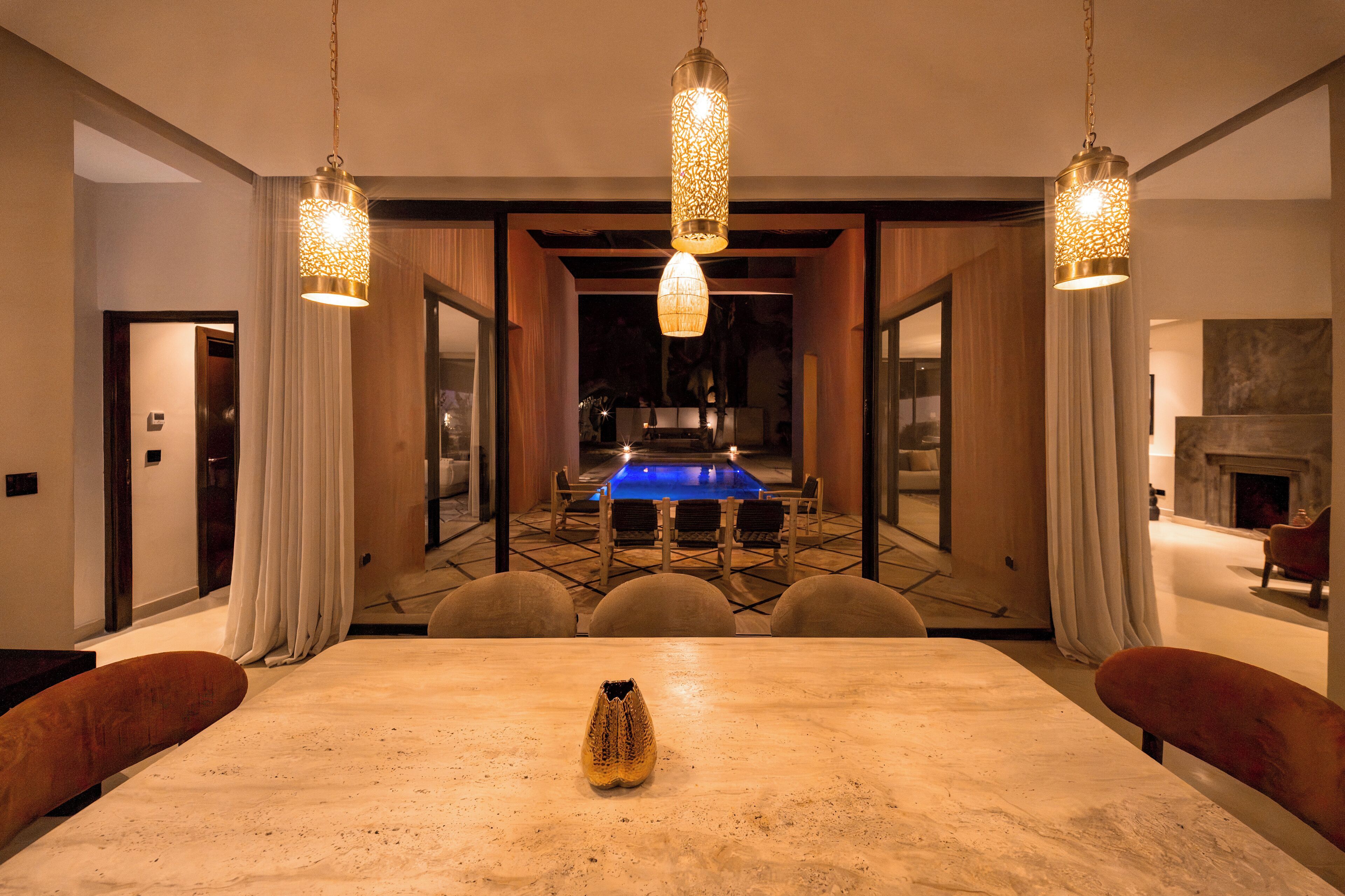 A modern dining room with a large table, hanging pendant lights, and a view of a pool and fireplace.