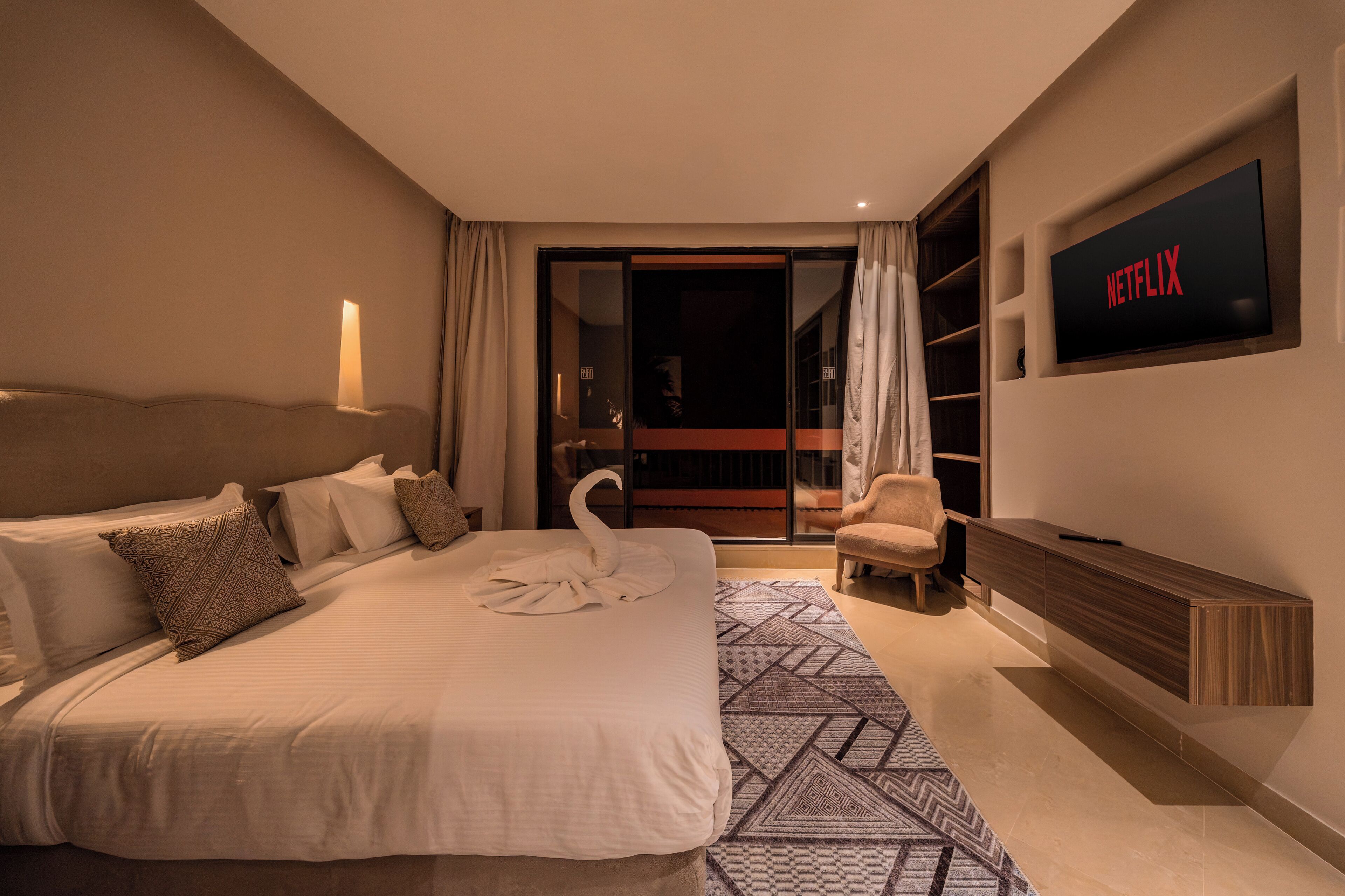 A hotel room with a bed, a TV mounted on the wall, a chair, and a sliding door leading to a balcony.
