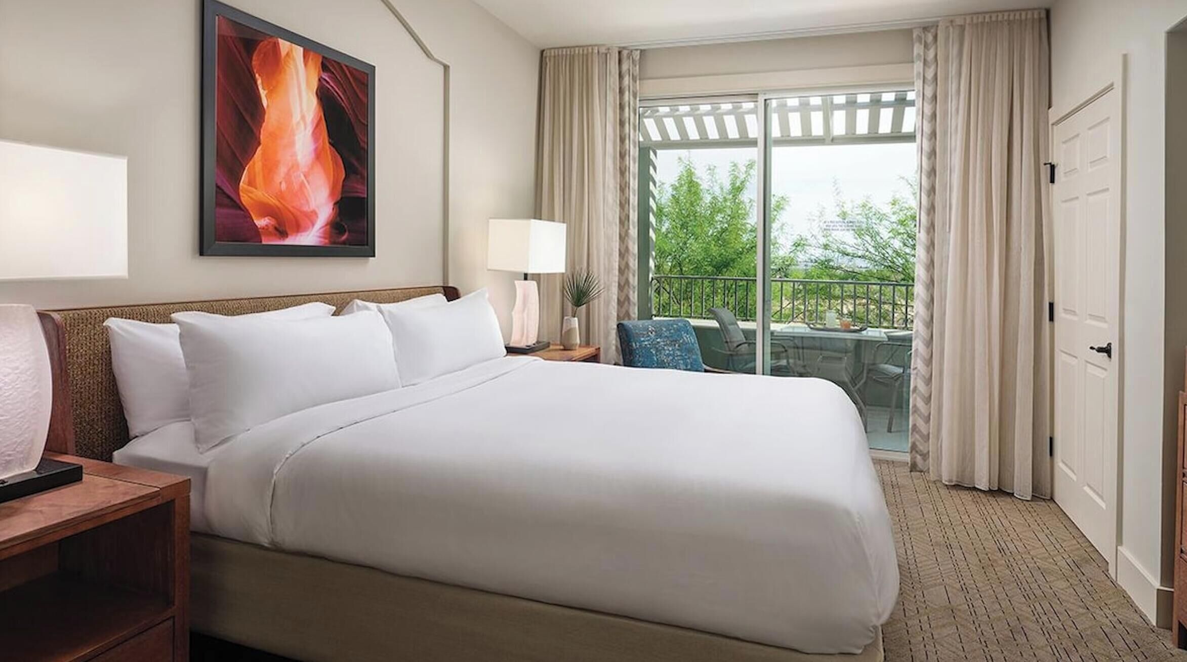 Une chambre d ’hôtel avec un grand lit, un bureau avec une lampe, une vue sur l’extérieur grâce à une porte coulissante, et un tableau accroché au mur.