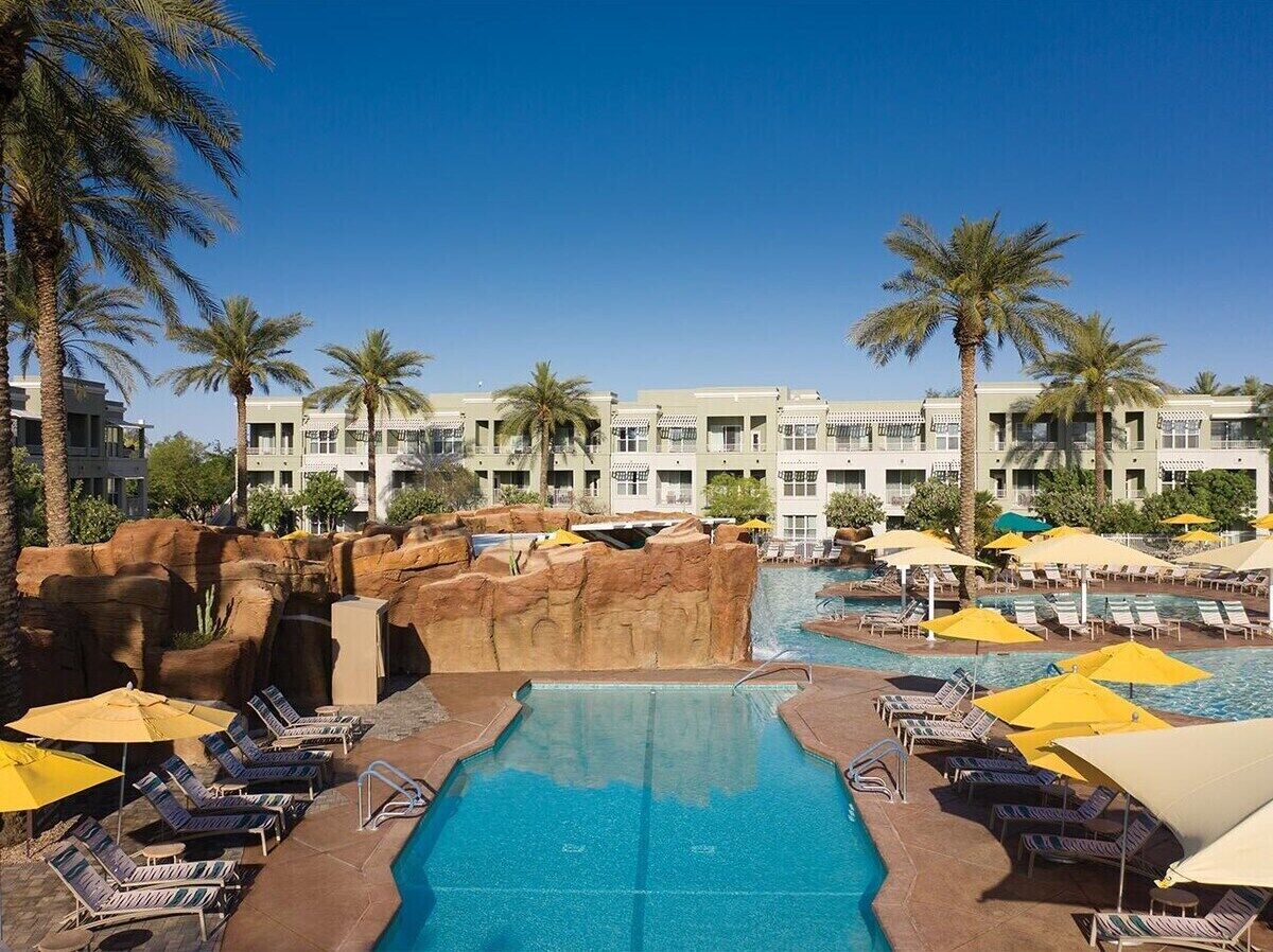 A hotel pool area with lounge chairs and umbrellas, surrounded by palm trees and a rocky landscape.