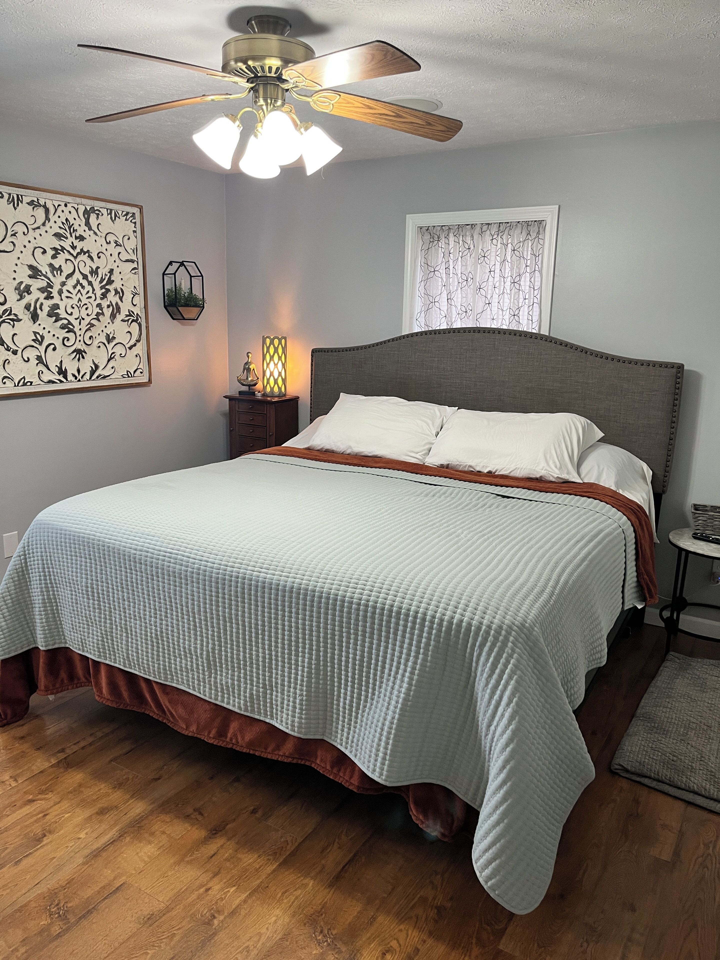 Une chambre à coucher comprenant un lit, un ventilateur de plafond, une table de chevet avec une lampe et une œuvre d’art accrochée au mur.
