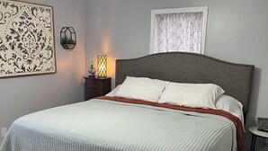 Un dormitorio con cama, ventilador de techo, mesita de noche con lámpara y una obra de arte en la pared.