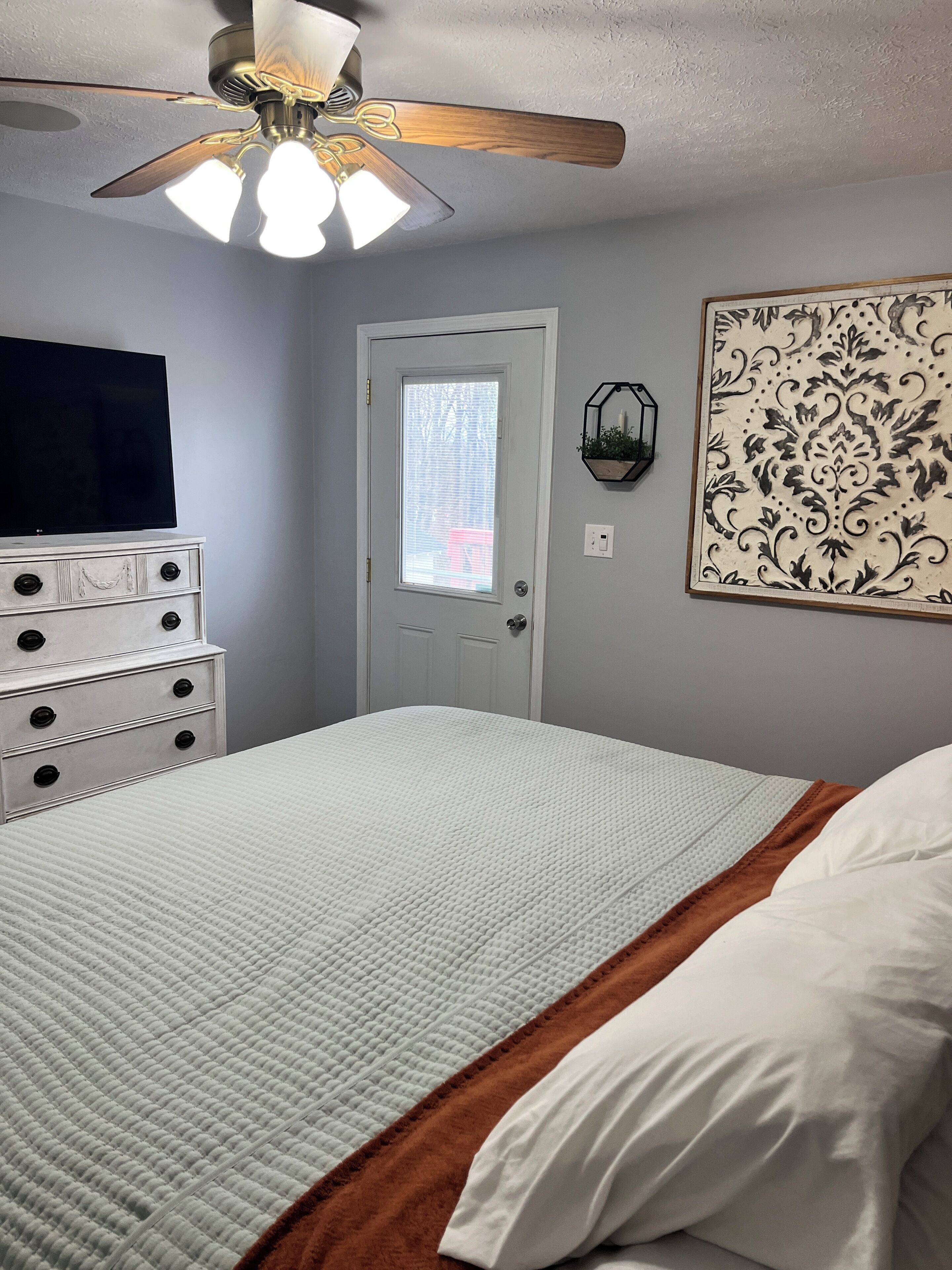 Una camera da letto con un letto, una cassettiera, una ventola a soffitto, una TV a muro e una porta con finestra.