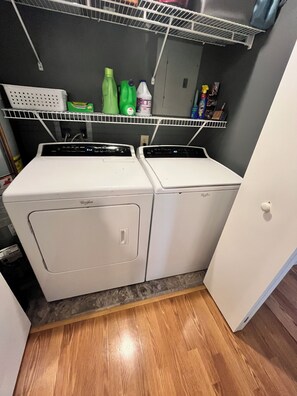 A laundry room with two stacked front-loading washers and dryers, a shelf above with cleaning supplies, and a wooden floor.
