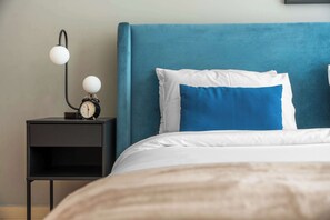 Ein Bett mit blauer Kopfteil, einem weiß-blauen Kissen, einem Nachttisch in Schwarz mit einer Uhr und einer Wandleuchte.