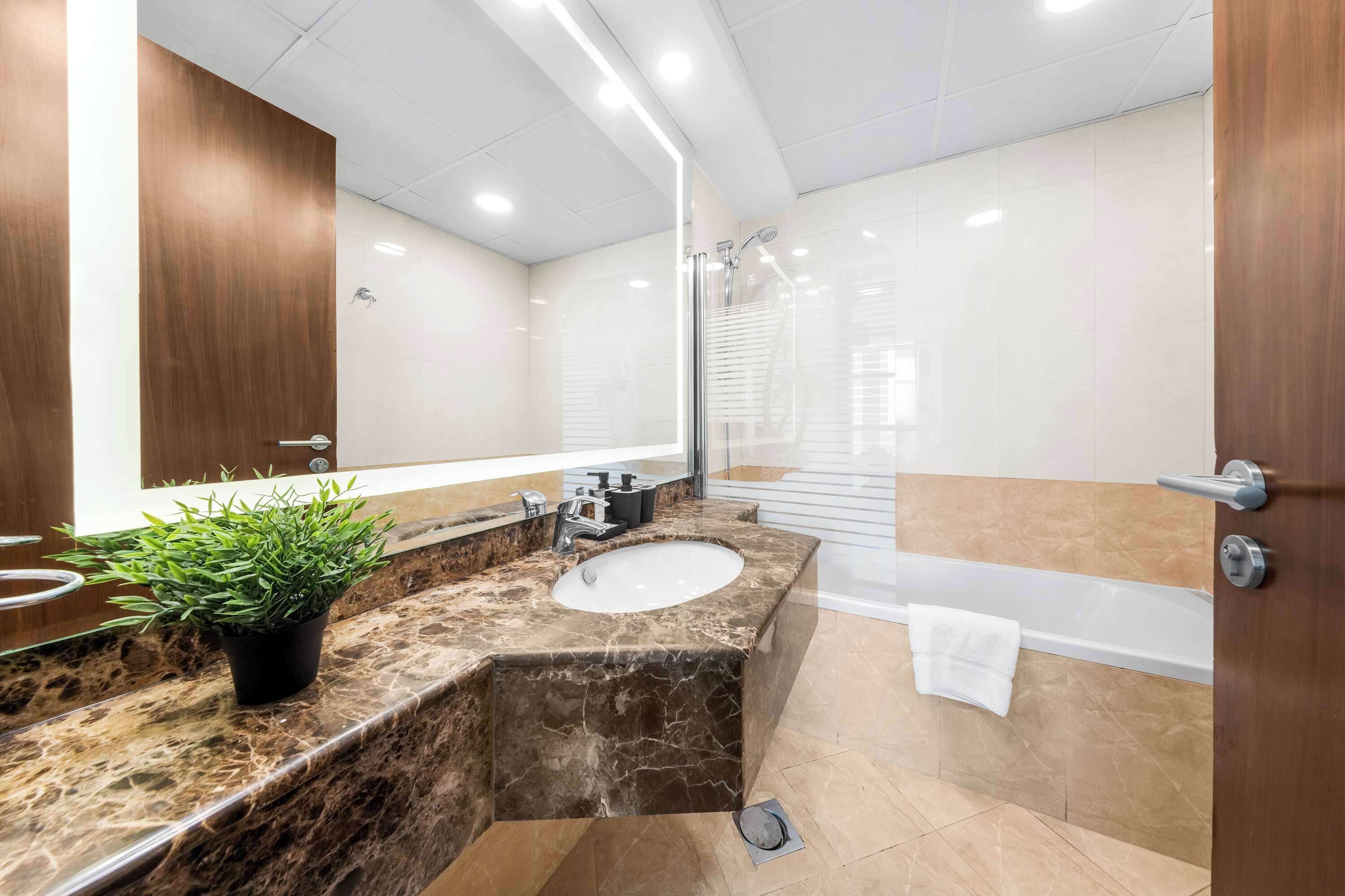A modern bathroom with a marble sink, a potted plant, a glass shower enclosure, and a white bathtub.