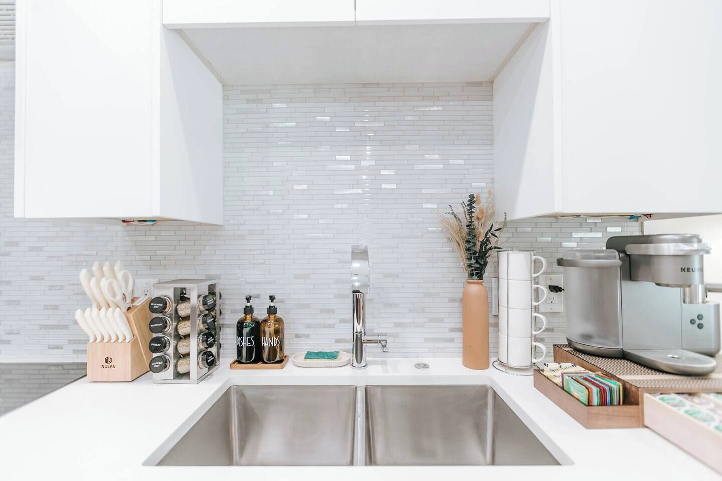 A modern kitchen with white cabinets, a stainless steel sink, a coffee machine, and a vase with dried plants.