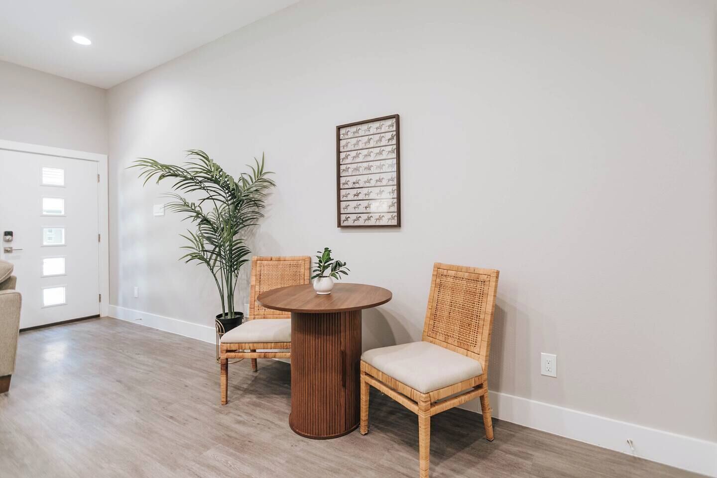 A room with a round wooden table, two wicker chairs, a potted plant, and a framed picture on the wall.