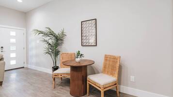 A room with a round wooden table, two wicker chairs, a potted plant, and a framed picture on the wall.