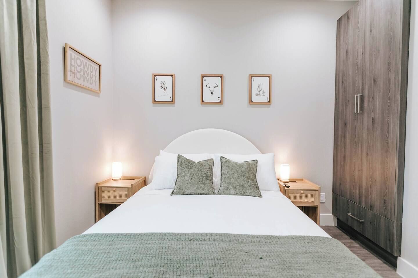 A bedroom with a white bed, two bedside tables with lamps, a wardrobe, and three framed pictures on the wall.