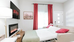 A hotel room with a bed, a TV mounted on the wall, a bedside table with a lamp, a window with red curtains, and a door. - Modern Condo Near Downtown Fort Worth & Stockyards (Fort Worth)