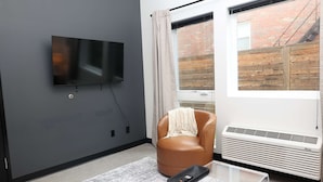 A modern living room with a flat-screen TV, a leather armchair, a glass coffee table, a window with a view of a brick wall, and a wall-mounted air conditioner.