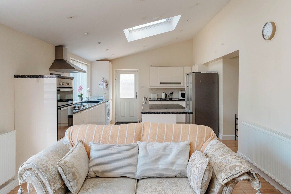A modern kitchen with a built-in oven, a refrigerator, and a microwave. There is a sofa with patterned cushions in the foreground.