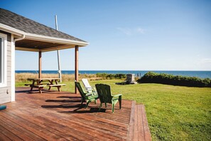 A wooden deck with a covered area, picnic table, and green chairs overlooking a grassy area with a view of the ocean.