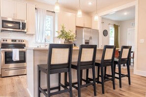 A modern kitchen with stainless steel appliances, a central island with bar stools, and a dining area with a view of the outdoors. - Raleigh Modern - Stylish 4BR - 5 TVs - Downtown (Raleigh)