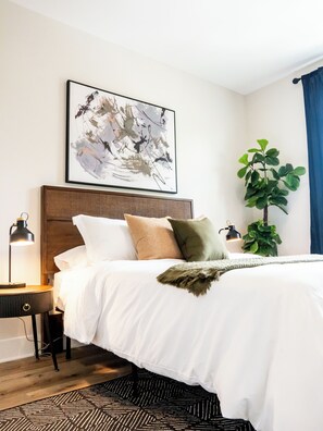 A neatly made bed with white bedding and a decorative throw, a wall-mounted abstract painting, a potted plant, and a bedside lamp. - Raleigh Modern - Stylish 4BR - 5 TVs - Downtown (Raleigh)