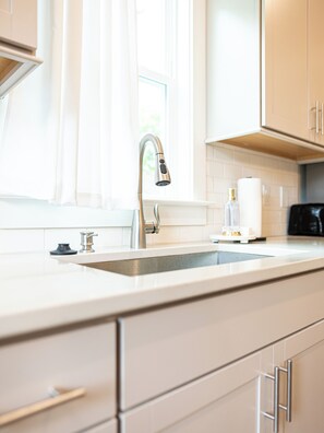 A modern kitchen with a stainless steel faucet, a white countertop, and a window with sheer curtains. - Raleigh Modern - Stylish 4BR - 5 TVs - Downtown (Raleigh)