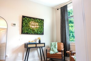 A room with a wall-mounted neon sign, a small table with a vase and books, a chair with a blanket, and a window with curtains. - Raleigh Modern - Stylish 4BR - 5 TVs - Downtown (Raleigh)