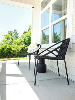 Two outdoor chairs with a woven backrest and a cushion with a face design, positioned on a concrete patio in front of a white building with large windows. - Raleigh Modern - Stylish 4BR - 5 TVs - Downtown (Raleigh)
