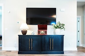 A dark blue cabinet with two doors, a flat-screen TV mounted on the wall above, a table lamp, a radio, and a vase with white flowers. - Raleigh Modern - Stylish 4BR - 5 TVs - Downtown (Raleigh)