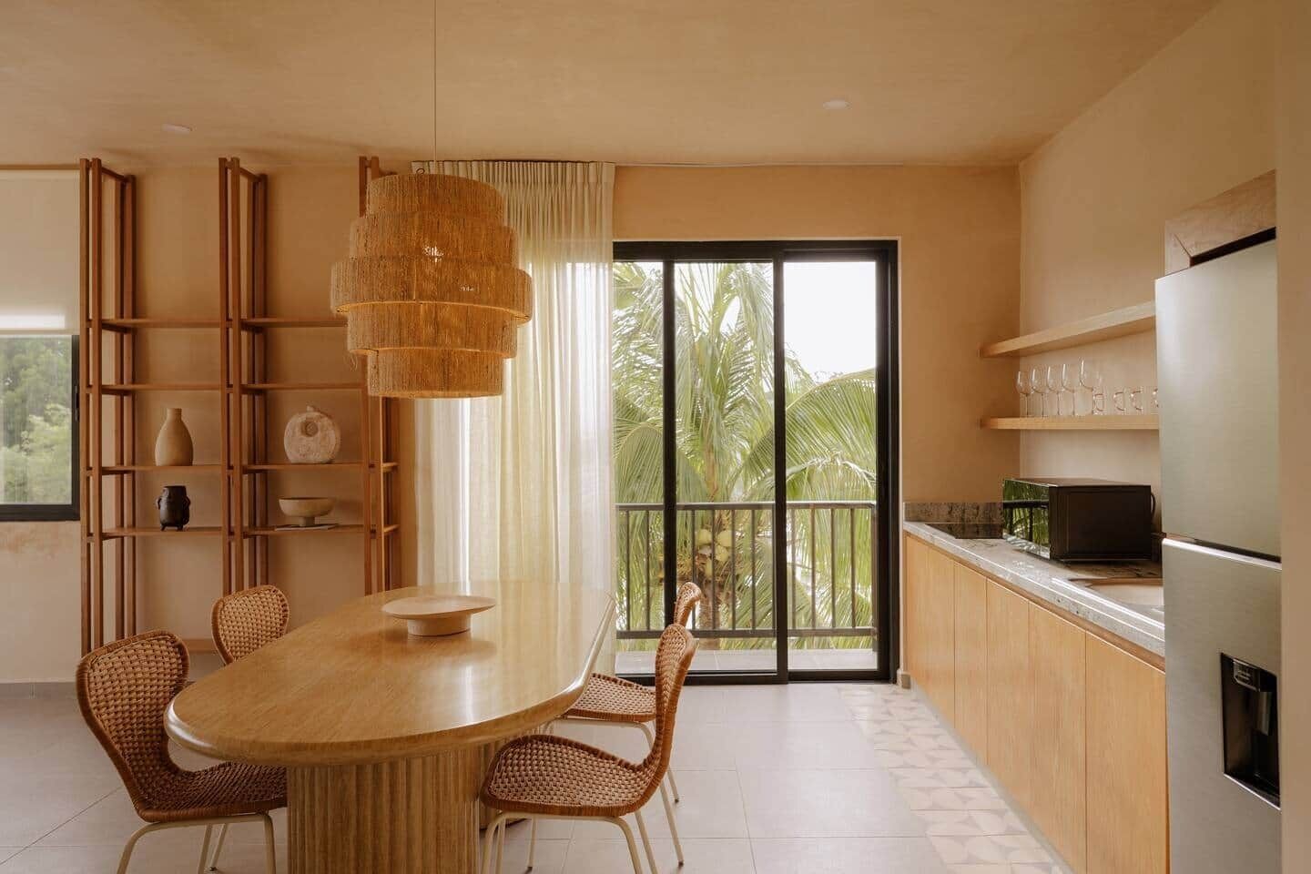 Una cocina moderna con mesa, sillas, lámpara colgante y vistas a palmeras a través de amplios ventanales.
