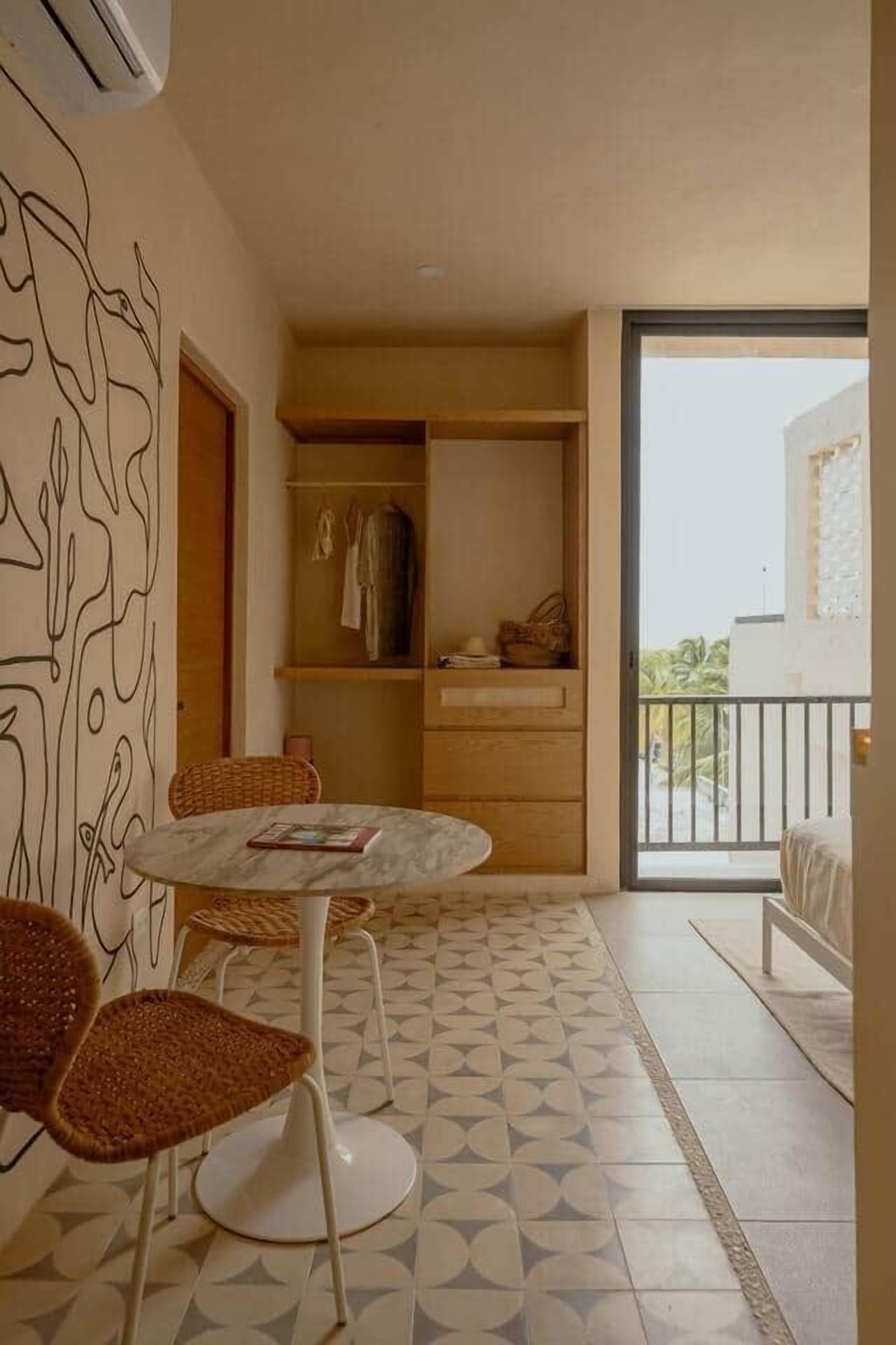 Une pièce avec une table ronde, deux chaises en osier, une armoire, une petite étagère avec des vêtements et, à travers une porte vitrée, une vue sur un balcon extérieur.