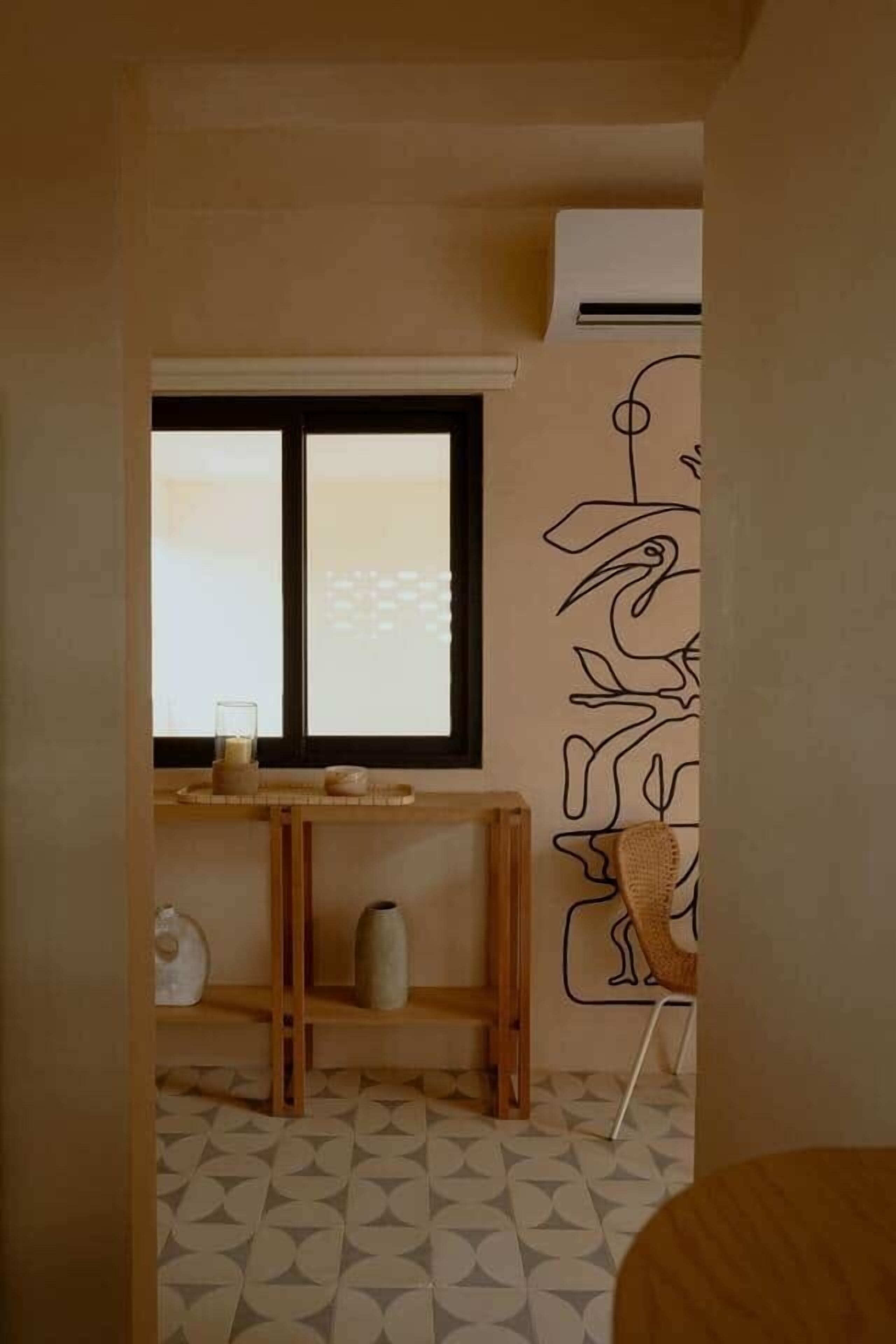 A room with a window, a wooden table with a vase, a chair, and a wall with a line drawing.