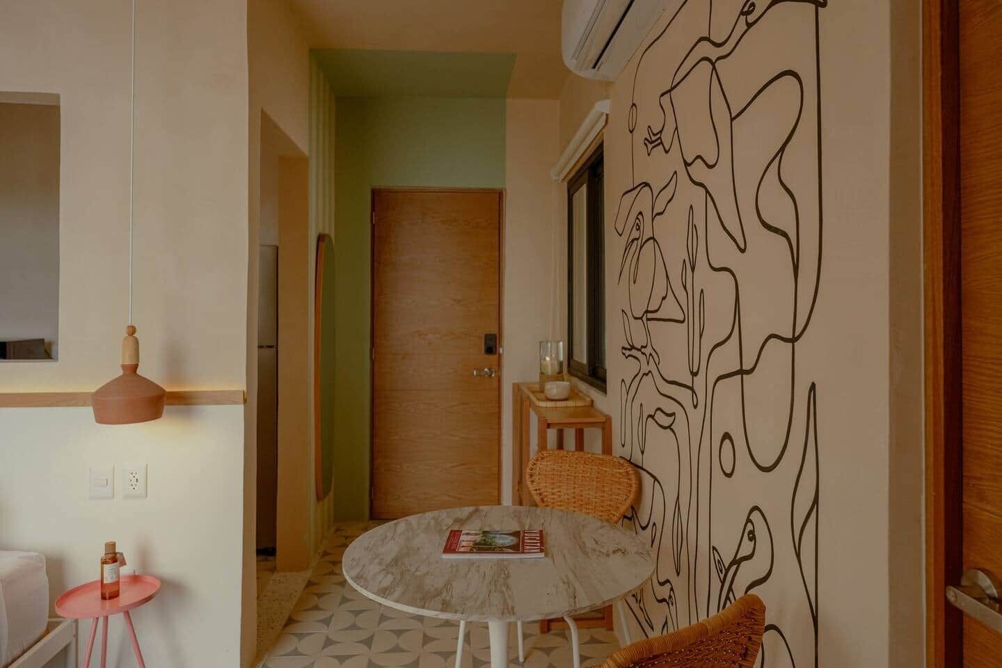 Una habitación con una mesa redonda, una silla, un taburete pequeño, una pared con un dibujo lineal en negro y una puerta al fondo.