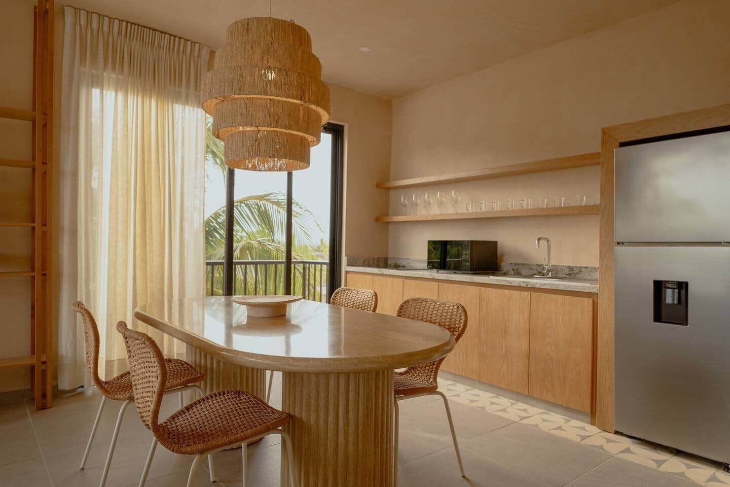 Une cuisine moderne avec un coin repas, comprenant une table ronde, des chaises en osier, un réfrigérateur en acier inoxydable et une grande fenêtre offrant une vue dégagée.