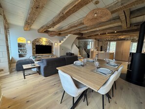 Un vaste espace de vie comprenant une table à manger en bois pour quatre personnes, une cheminée en pierre et, au fond, une cuisine moderne.