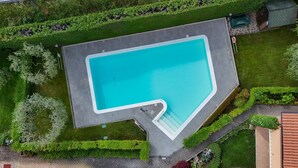 Ovenudblik over en swimmingpool med et unikt L-formet design, omgivet af velplejede græsplæner og hække.