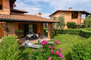 A patio with a table and chairs, a small garden with flowers, and a building with a tiled roof. - MGH - Garda Bloom Family Stay Manerba (Manerba del Garda)
