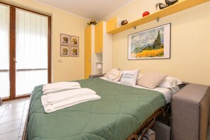A hotel room with a bed, a chair, a small table, a painting, and a door with a view. - MGH - Garda Bloom Family Stay Manerba (Manerba del Garda)