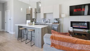 A modern kitchen with a central island, bar stools, a dining area, a living room with a sofa, a fireplace, and a TV mounted on the wall. - Stockyards + Downtown | Bright 1BR | Sleeps 3 - Apartment (Fort Worth)