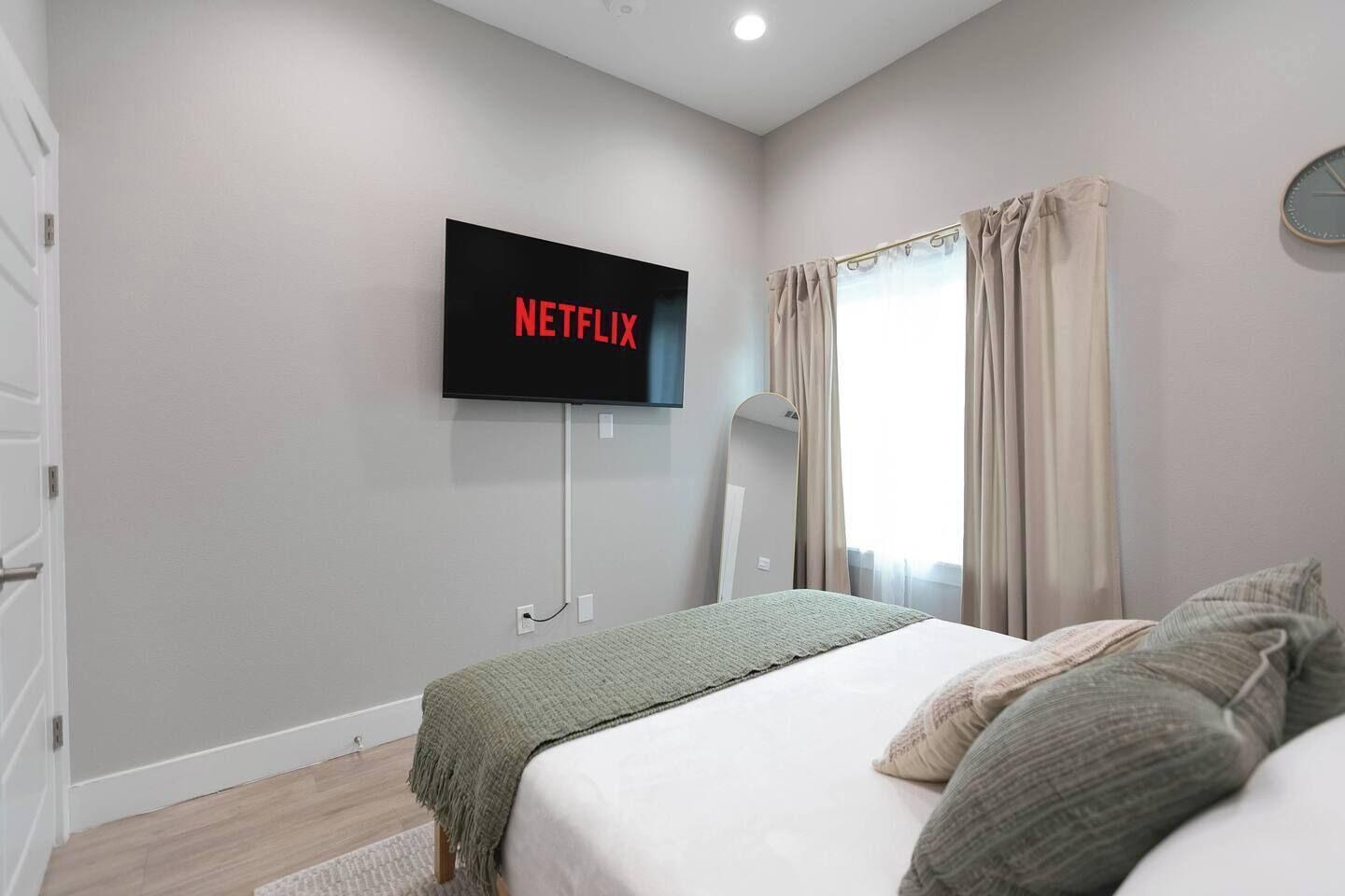 A bedroom with a bed, a wall-mounted TV displaying 'NETFLIX,' a mirror, and a window with curtains.