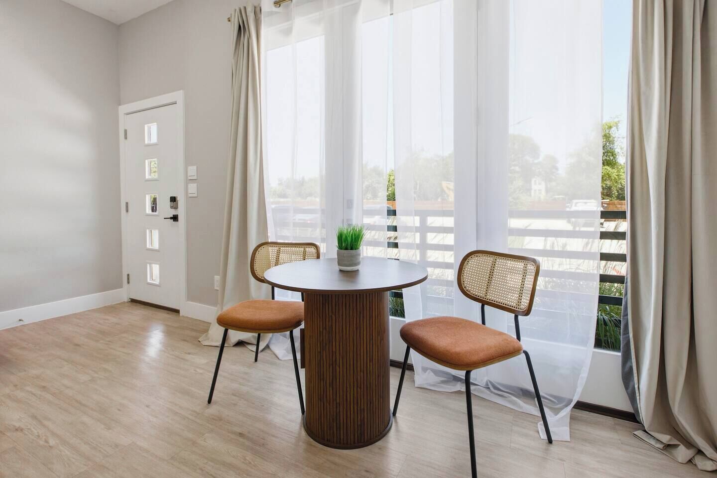 A room with a round wooden table, two wicker chairs, a potted plant, and a view through sheer curtains.