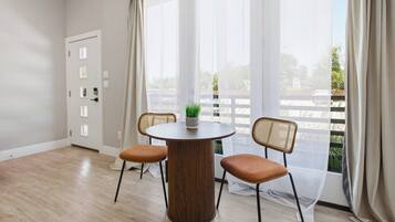 A room with a round wooden table, two wicker chairs, a potted plant, and a view through sheer curtains.