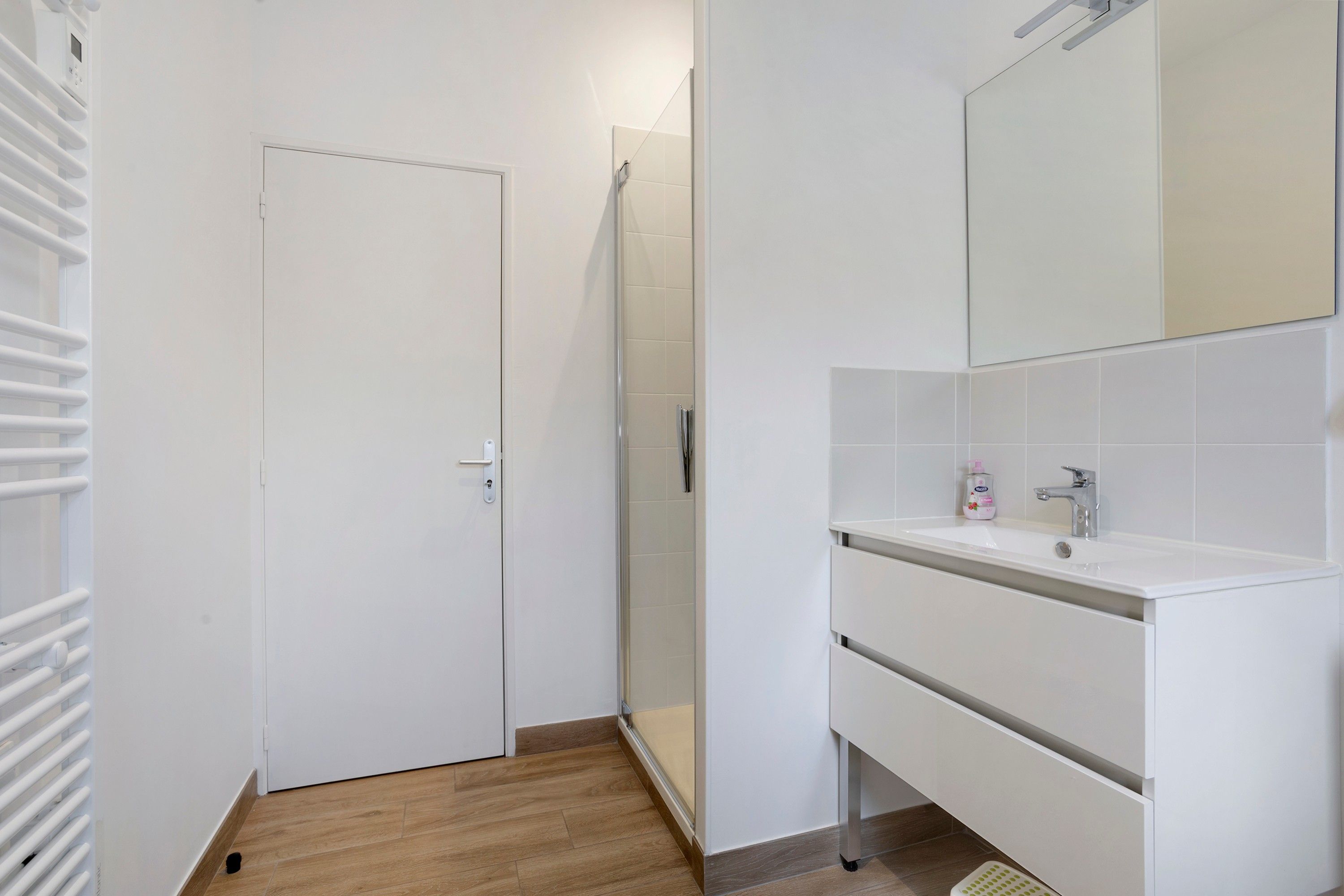 Ein Badezimmer mit einer weißen Tür, einer Duschkabine, einem Waschbecken mit Spiegel und einer Seifenflasche.