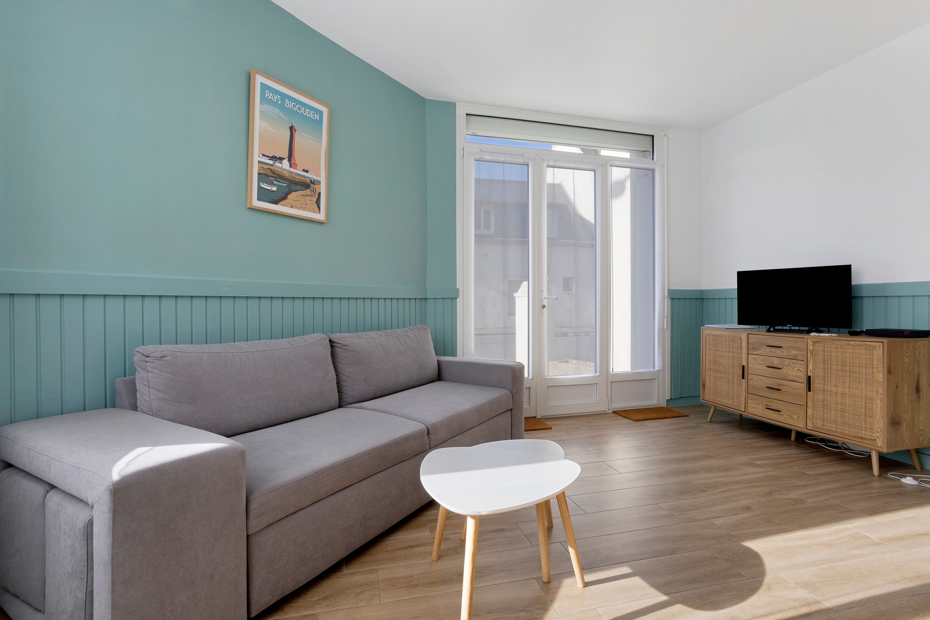 Una sala de estar con un sofá gris, una mesita blanca, un mueble de madera con un televisor y un ventanal con vista a edificios.
