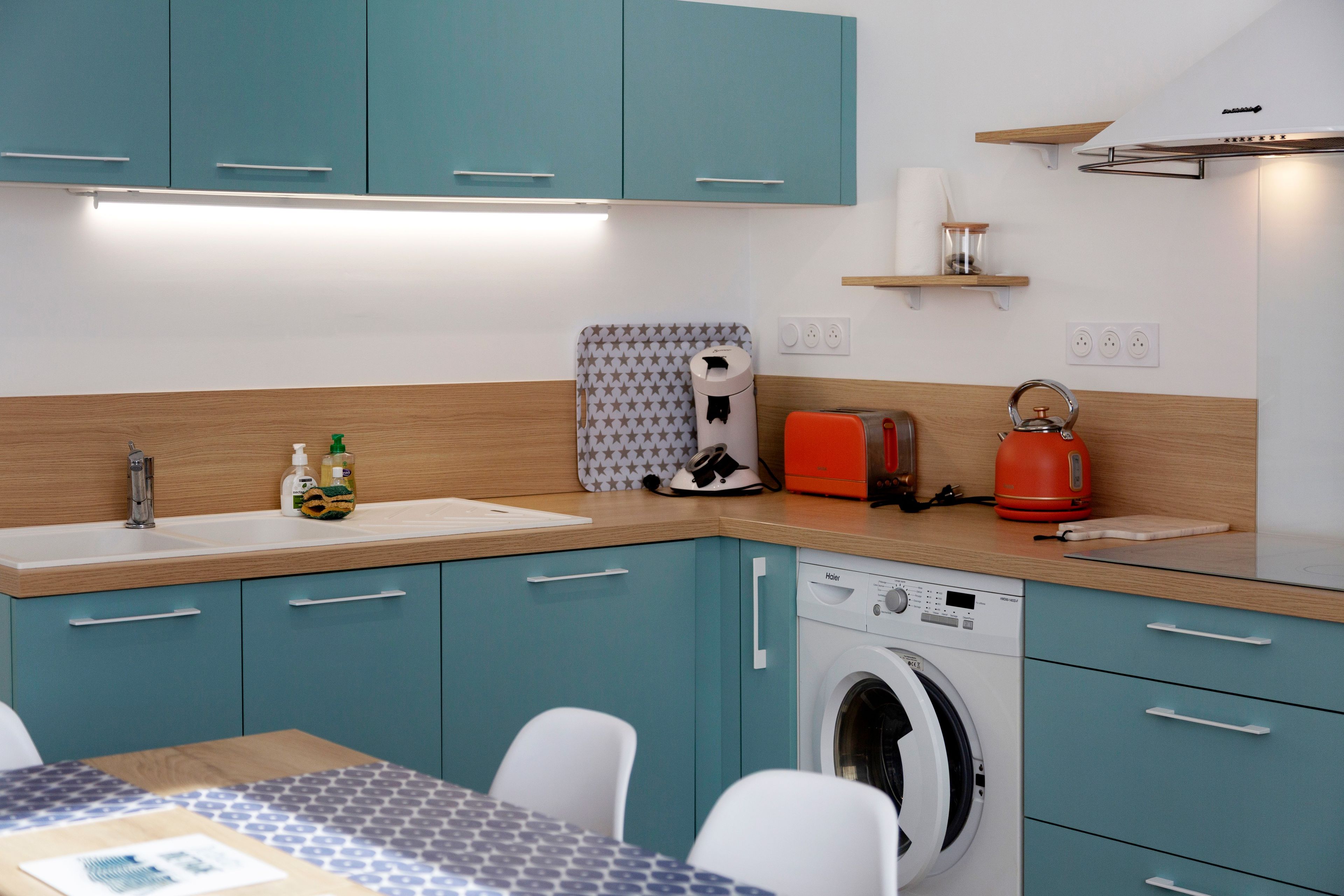 Een compacte keuken met blauwgroene kastjes, een ingebouwde wasmachine, een koffiezetapparaat en een waterkoker op het aanrecht.
