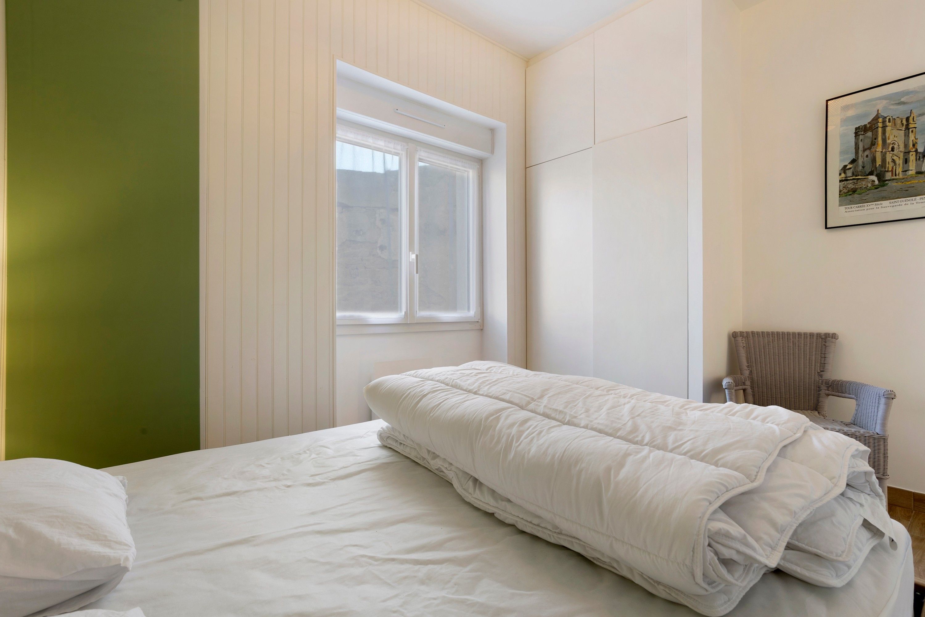 A single bed with a white duvet in a room with a green accent wall, a window, and a framed picture on the wall.