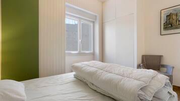 A single bed with a white duvet in a room with a green accent wall, a window, and a framed picture on the wall.
