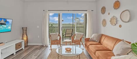 A living room with a sofa, coffee table, and chairs, a TV, and a view through a sliding door.
