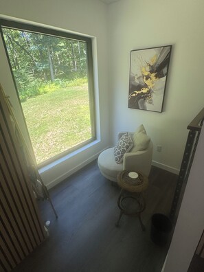 Ein Zimmer mit großem Fenster, einem Sessel mit Kissen, einem kleinen runden Tisch und einem an der Wand hängenden abstrakten Gemälde.