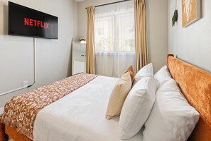 Ein Hotelzimmer mit Bett, Wandfernseher, Fenster mit Vorhängen und Nachttisch.