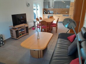 Ein Wohnzimmer mit Fernseher, Holztisch, Stühlen, einer Couch und einer Küche im Hintergrund.