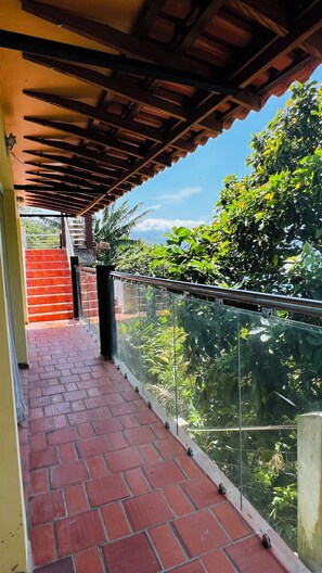Un balcón techado con suelo de ladrillo, barandilla de vidrio y vistas a la vegetación.