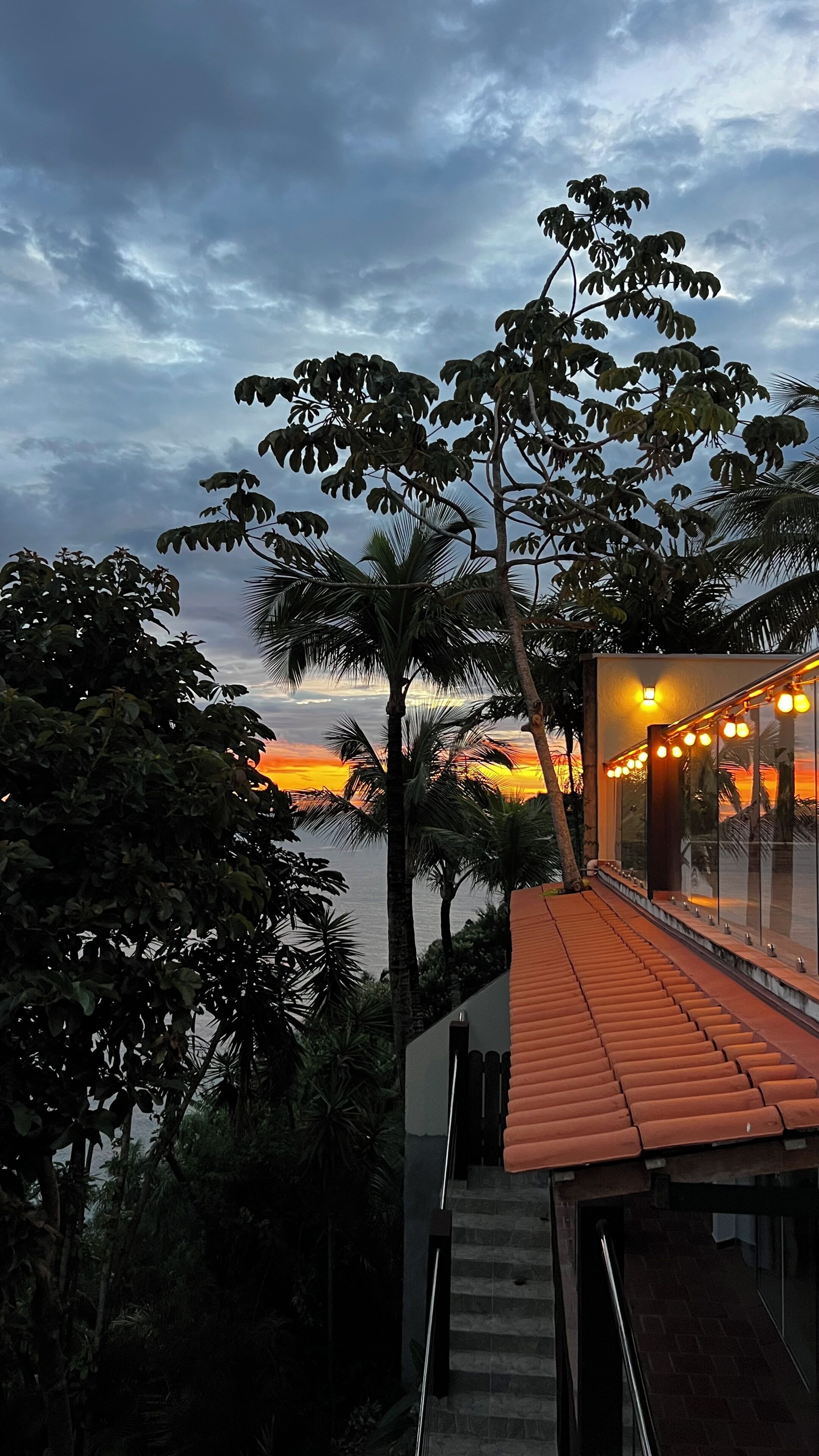 A sunset view from a balcony with a railing, palm trees, and a building with a tiled roof.