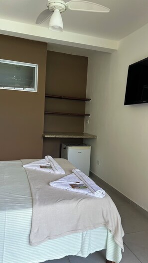 Habitación de hotel con cama, ventilador de techo, refrigerador pequeño, televisor de pantalla plana y ventana.